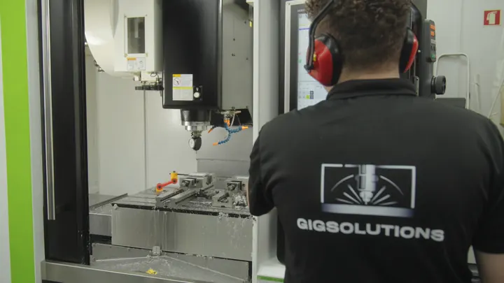Operador da GIG Solutions, de costas, com o polo da empresa e protetores auriculares, diante de um centro de maquinação CNC aberto