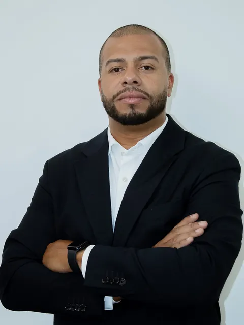 Ivan Lúcio, de blazer escuro e camisa branca, de braços cruzados, diante de uma parede clara